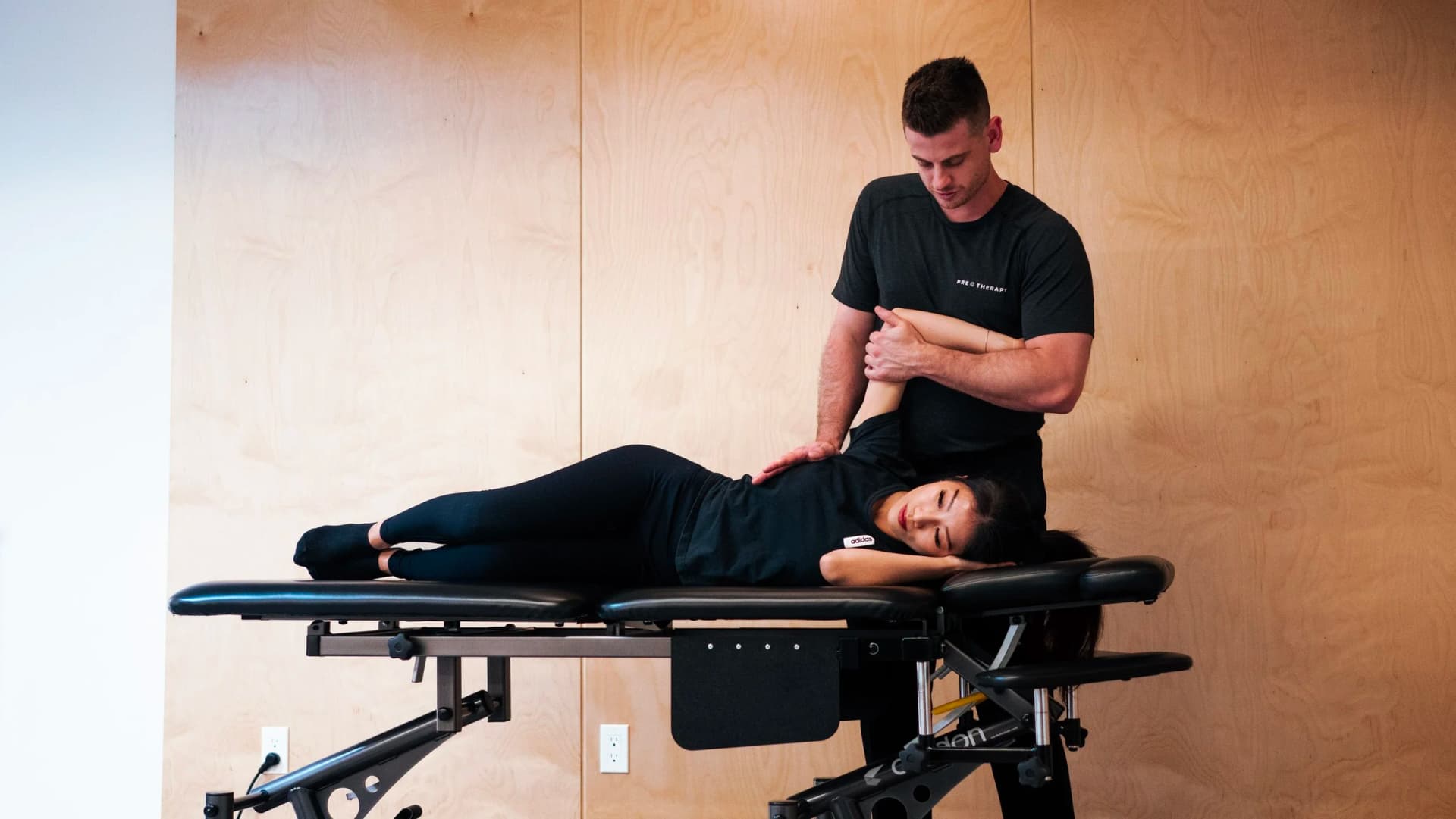 A therapist stretches a patient's arm behind her back to release her shoulder as she lies on her side on a treatment table.