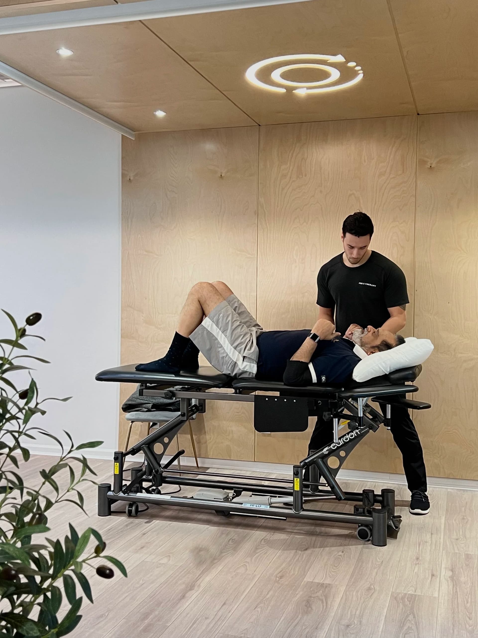 A therapist stands at the head of a treatment table supporting an older man's neck and shoulders as he lies with his knees bent.