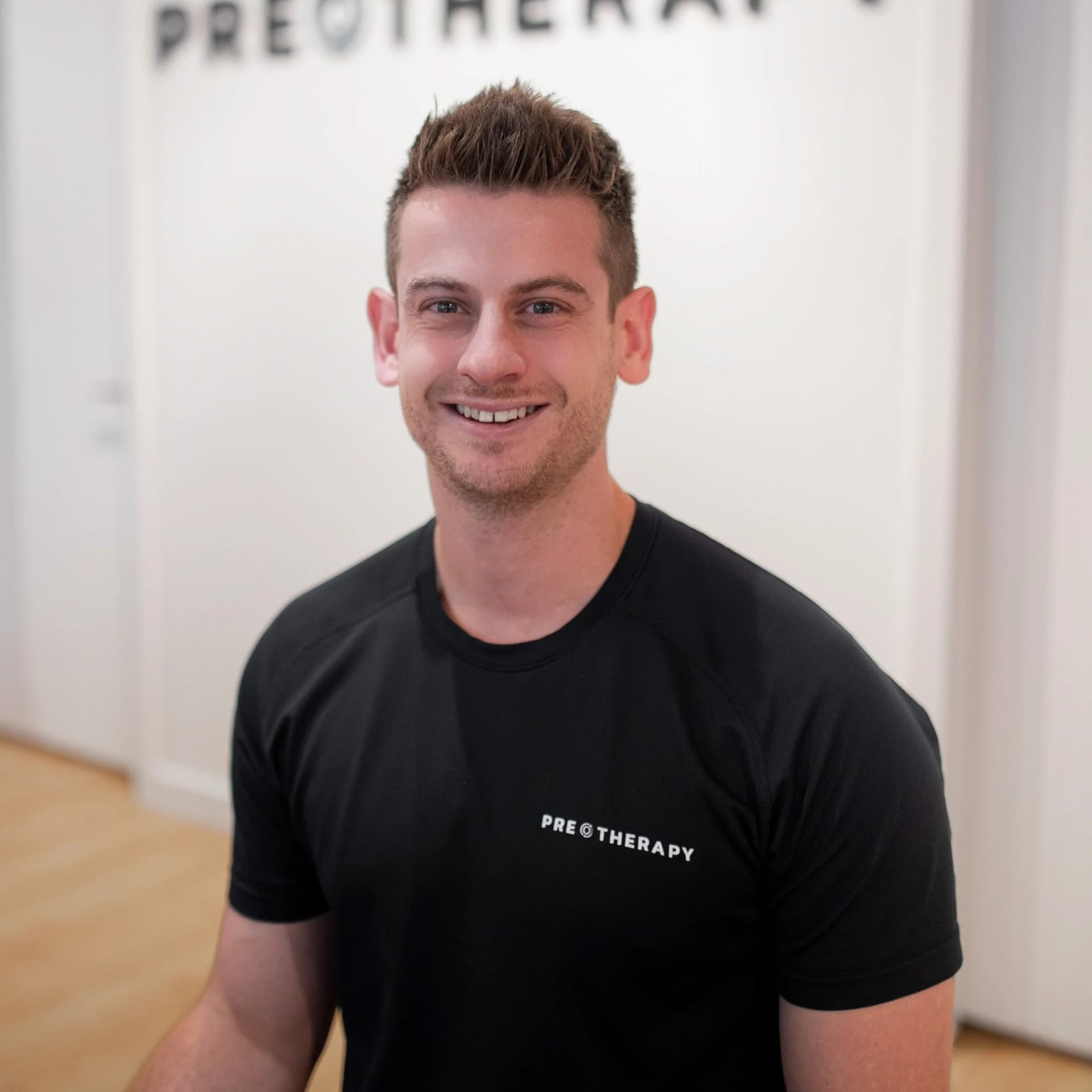A smiling practitioner in a black PRE Therapy shirt stands in front of the clinic's logo wall.