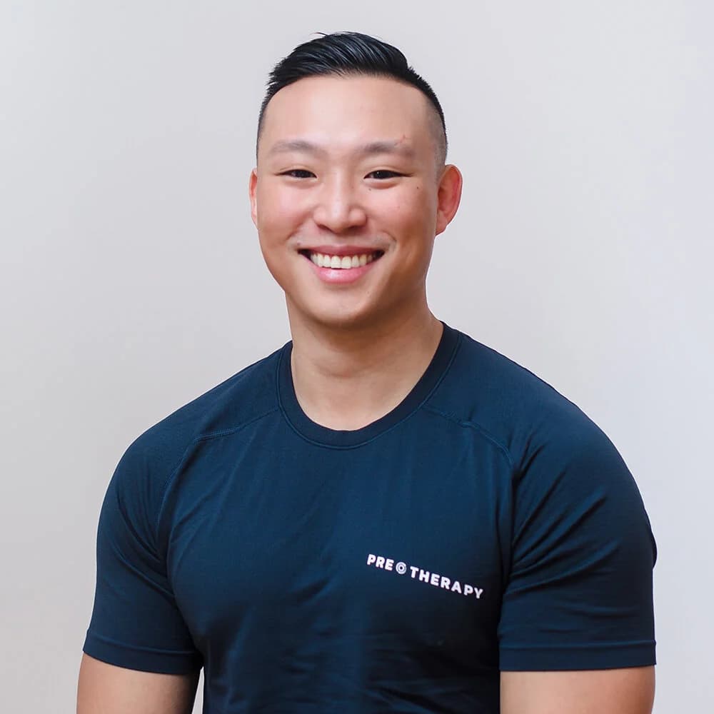 A smiling therapist in a navy PRE Therapy shirt poses for a headshot against a plain light backdrop.