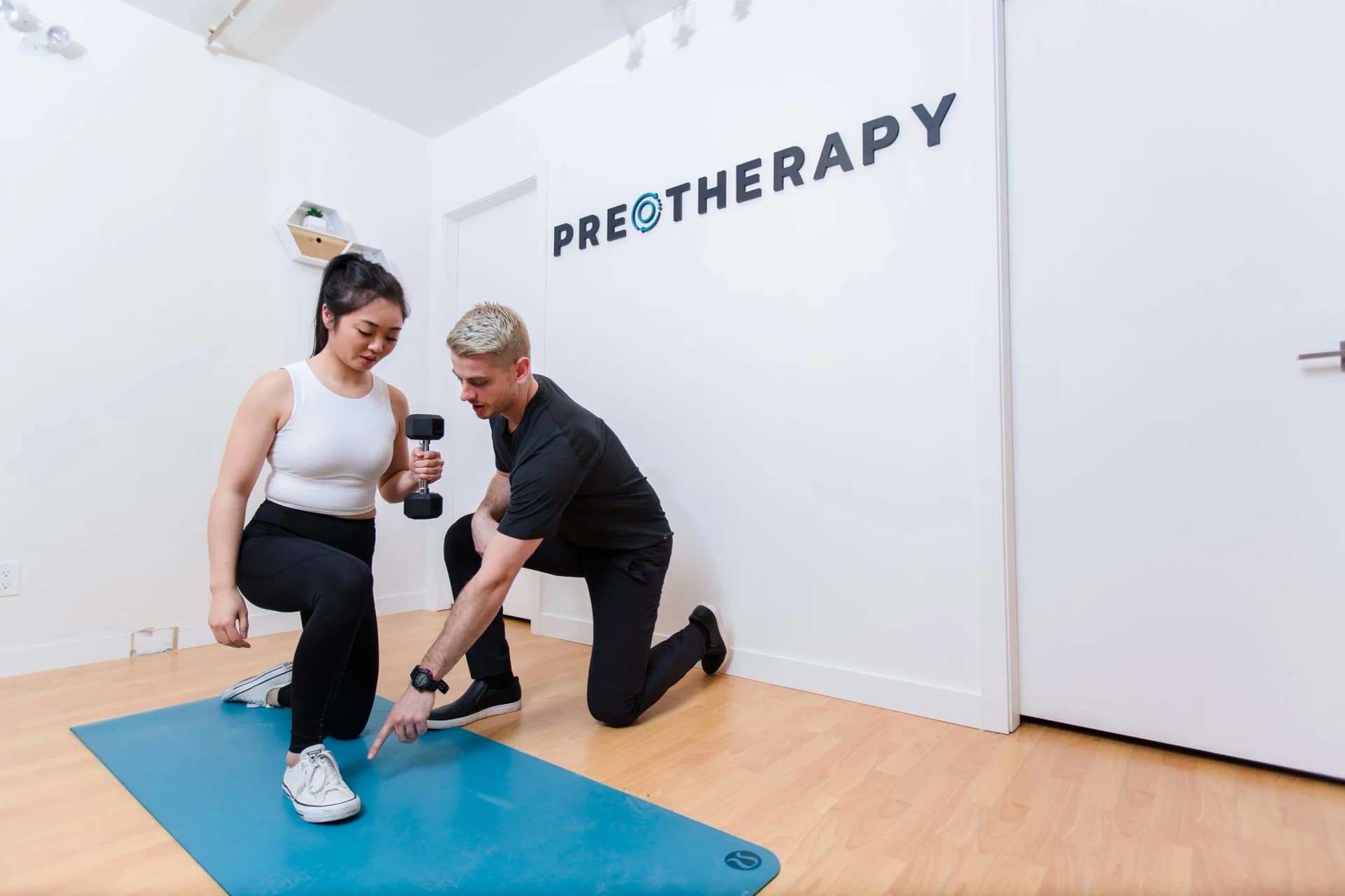 A kinesiologist points out foot placement as a client holds a dumbbell in a kneeling lunge on an exercise mat at PRE Therapy.