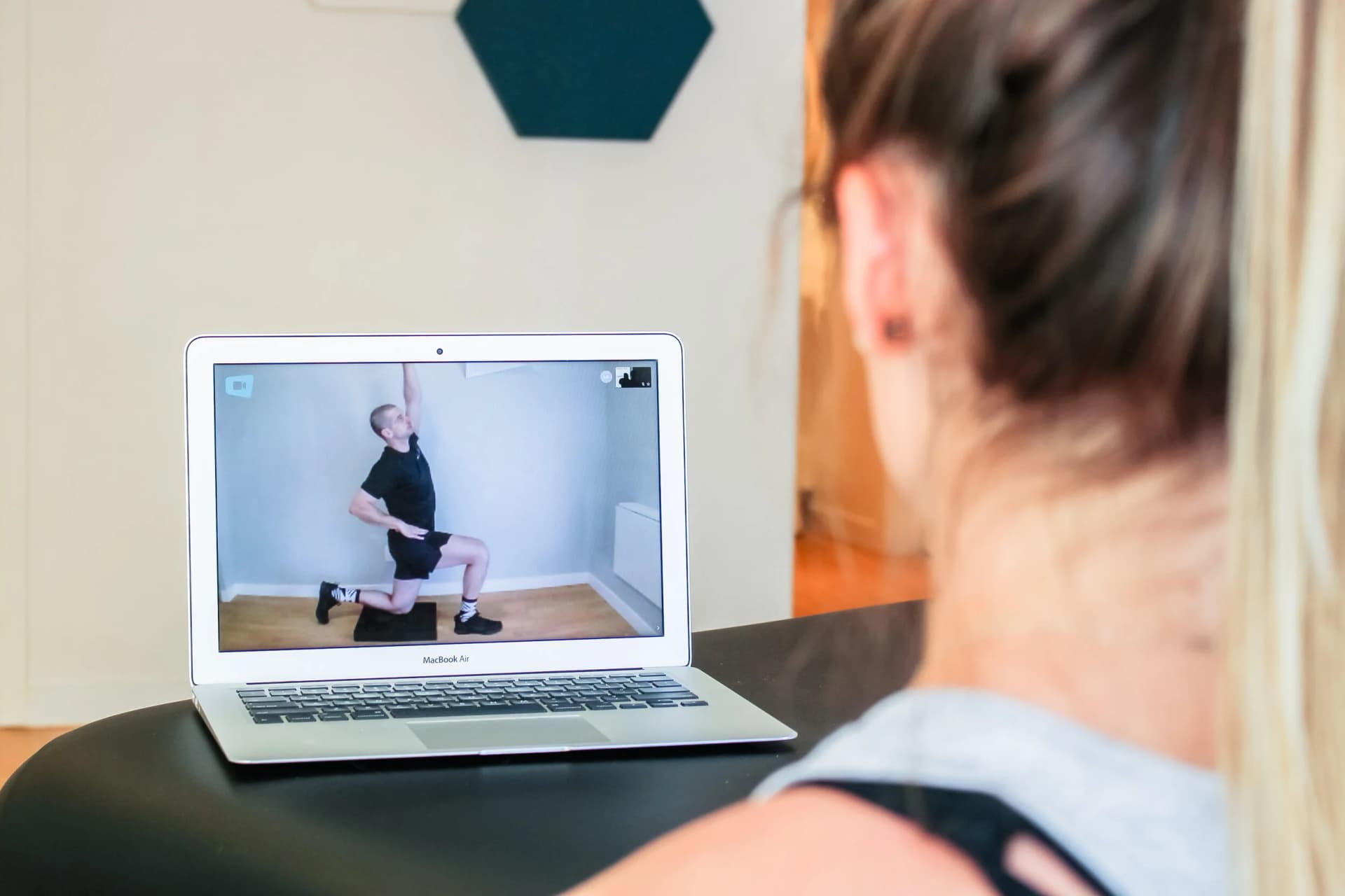 A woman watches a laptop where a kinesiologist demonstrates a half-kneeling reach exercise during a virtual therapy session.